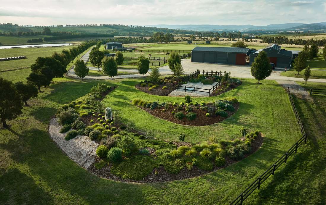 Aerial view of sweeping native garden beds curving through lawn toward the stables and sheds