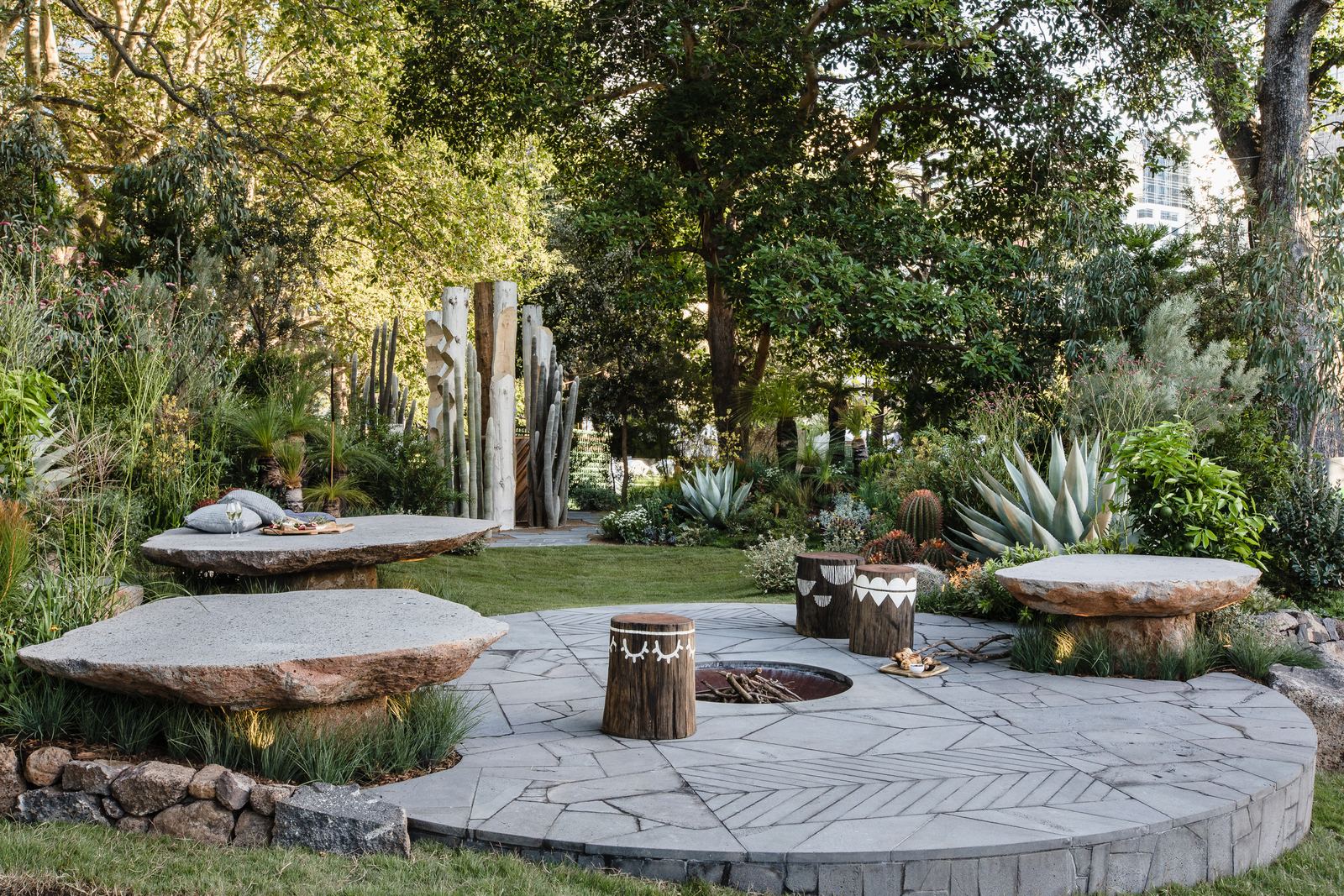 Stone monoliths and a sunken fire pit set in crazy paving, carved timber poles standing beyond the lawn