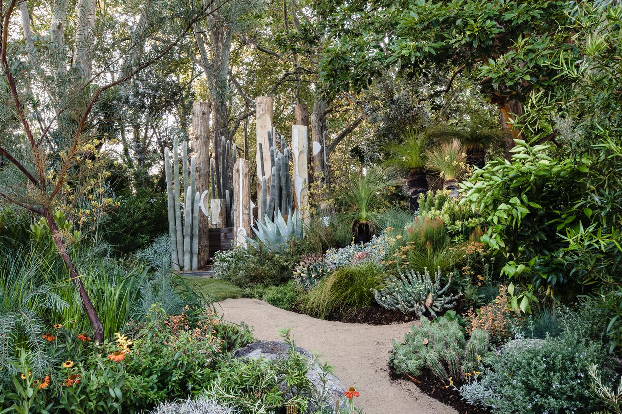 Decomposed granite path curving through dense planting toward the carved timber poles