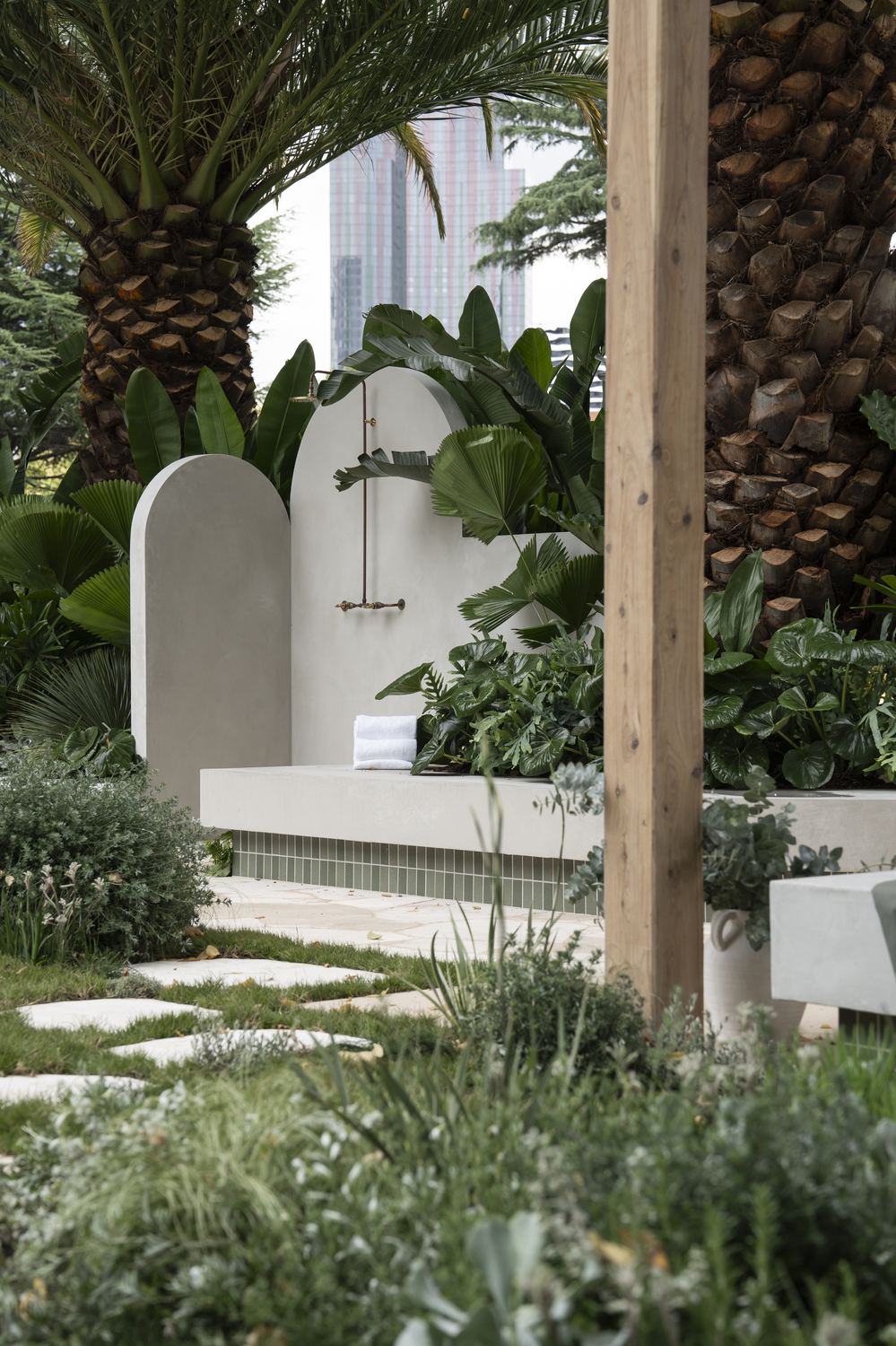 Two arched render walls carrying the brass shower, framed by fan palms and strelitzia