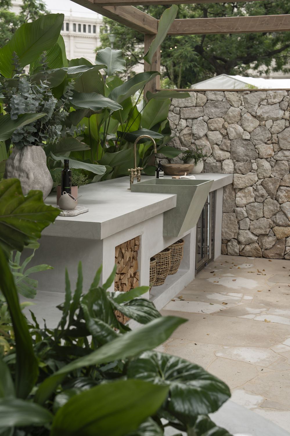 Concrete outdoor kitchen with a green trough sink, brass tapware and firewood stacked below
