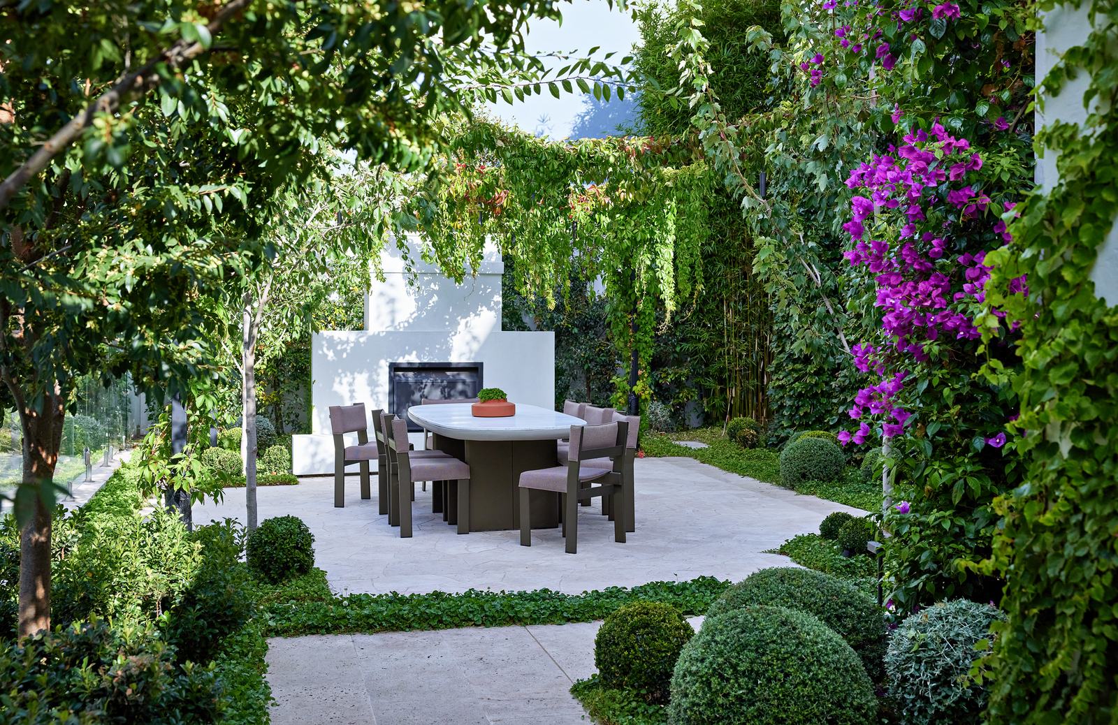 The dining terrace at Ormond Esplanade, an oval marble table set below a rendered outdoor fireplace under a canopy of ornamental grape