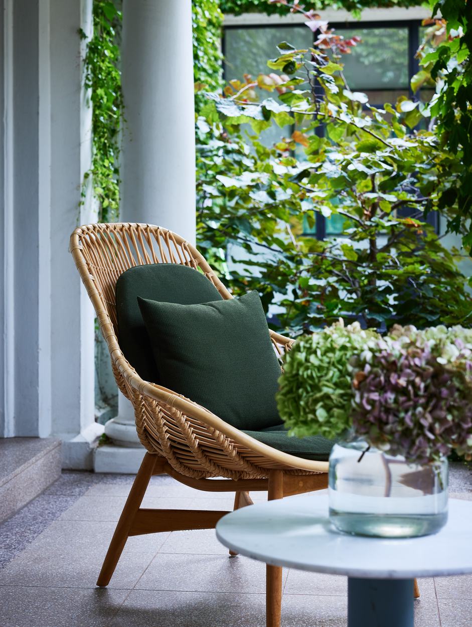 A cane armchair on the terrazzo loggia floor beside cut hydrangeas in a glass vase