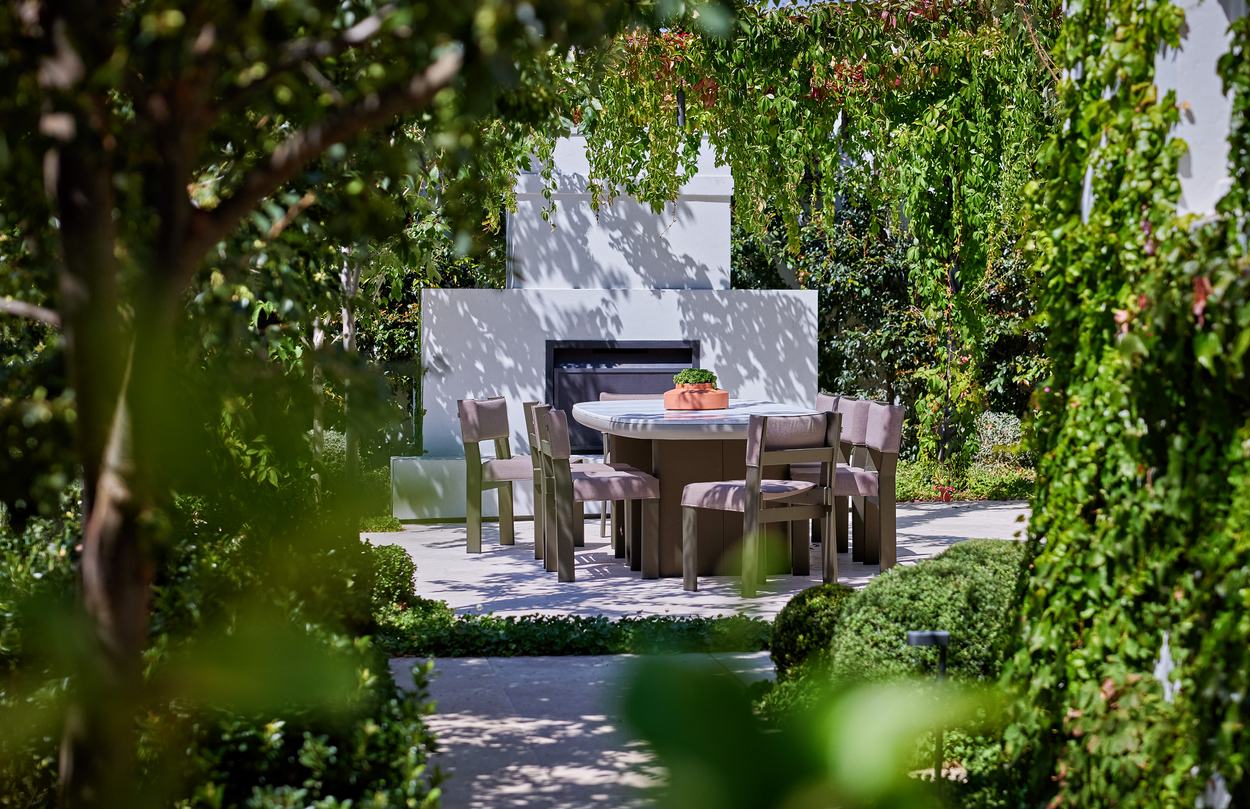 The dining table and fireplace glimpsed through the surrounding foliage, dappled light across the paving