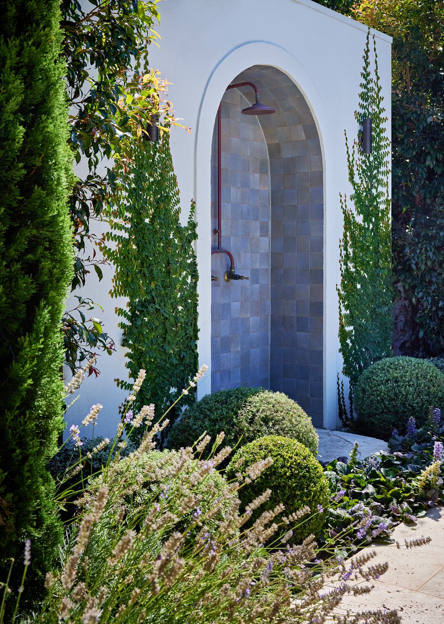 The arched shower recess lined in glazed blue tile with aged brass fittings, creeping fig running up the render either side